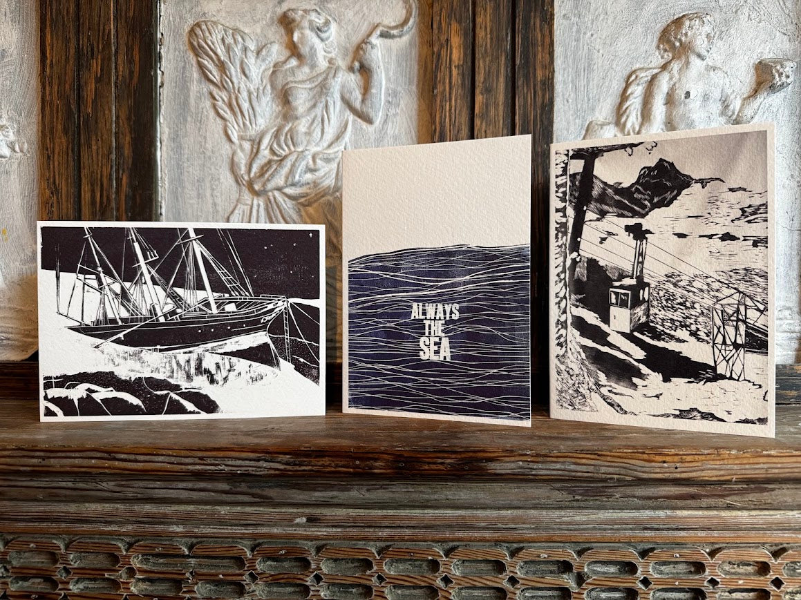 Three nautical-themed art prints displayed side-by-side on a rustic wooden mantelpiece. The prints feature black and white illustrations of a sailing ship, ocean waves with the text 'ALWAYS THE SEA', and a coastal lighthouse scene.