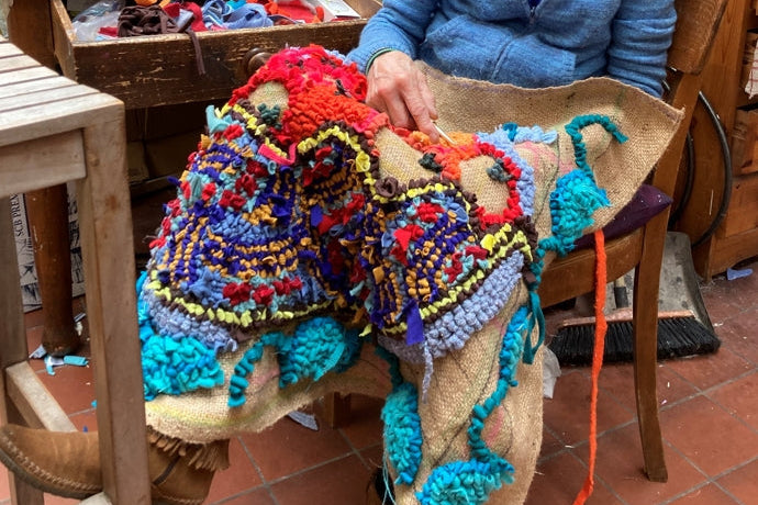 Artist and rug maker Lu Mason sitting in a workshop with a variety of colorful rug materials and tools.
