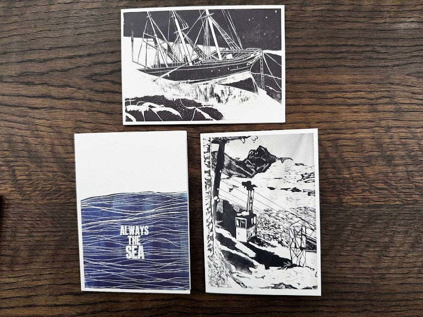 Three printed art cards or postcards arranged on a dark wood surface. Two cards show black-and-white abstract architectural or industrial line drawings, and the third card below shows a blue-and-white abstract pattern with the text 'AER FILM'.