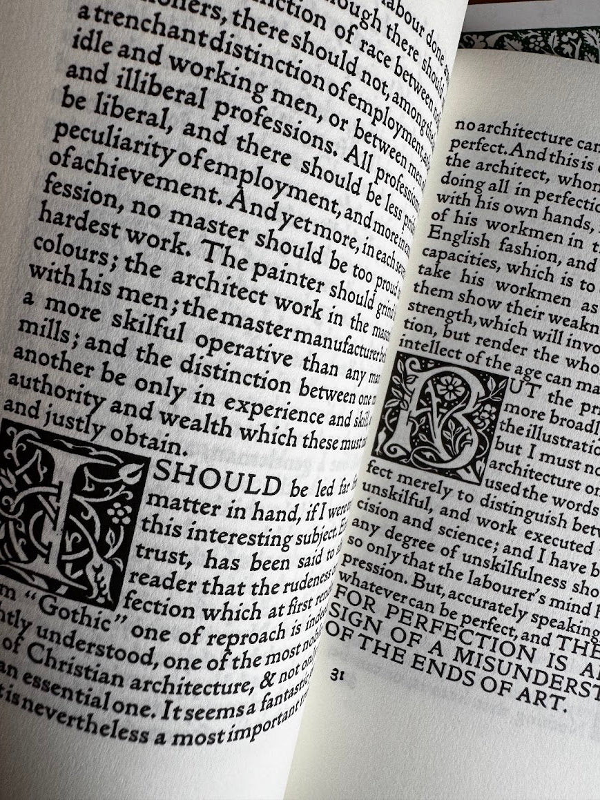 An open vintage book with dense black text on aged off-white pages, featuring an ornate decorative initial letter and floral border illustration.