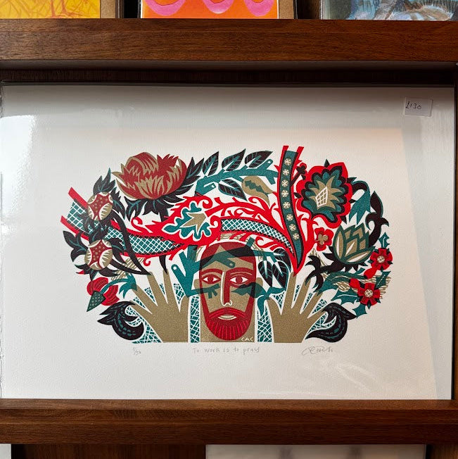 A framed art print featuring a stylized face surrounded by bold red, green, black, and beige floral and leaf motifs, displayed on a wooden shelf above other colorful artwork.