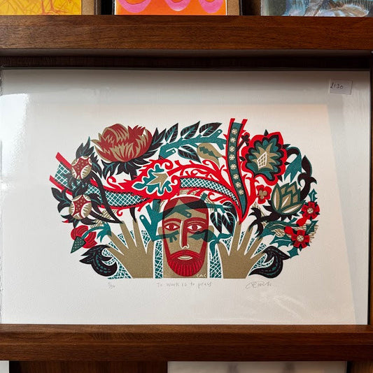 A framed art print featuring a stylized face surrounded by bold red, green, black, and beige floral and leaf motifs, displayed on a wooden shelf above other colorful artwork.